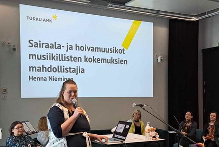 Henna Nieminen pitää seminaarissa puheenvuoroa ”Sairaala- ja hoivamuusikot musiikillisten kokemuksien mahdollistajia”, teksti näkyy taustalla dialla.
