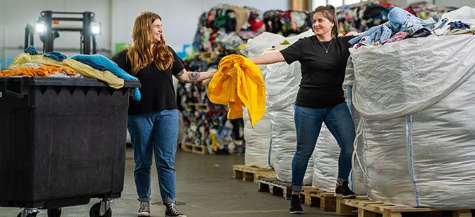 Kaksi nuorta naista seisoo kuvassa keskellä tekstiilien kierrätystä.