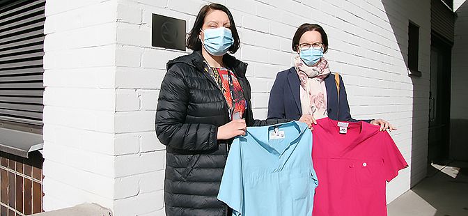 Koulutus- ja tutkimuspäällikkö Virpi Fagerström ja Naantalin kaupungin suun terveydenhuollon osastonhoitaja Kirsi Koivisto seisovat vierekkäin maskit kasvoillaan ja esittelevät lahjoitettuja suojavaatteita, punaista ja sinistä paitaa.