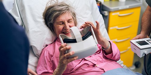 Vanhempi rouva vaaleanpunaisessa sairaalapyjamassa tutkii sairaalasängyllä vr-laseja käsissään.