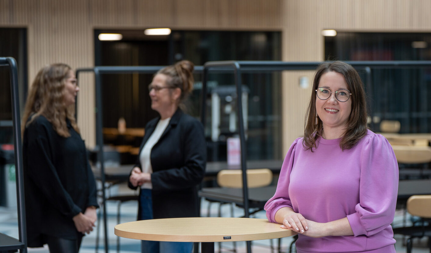 Henkilö seisoo pöydän ääressä ja katsoo hymyillen kameraan