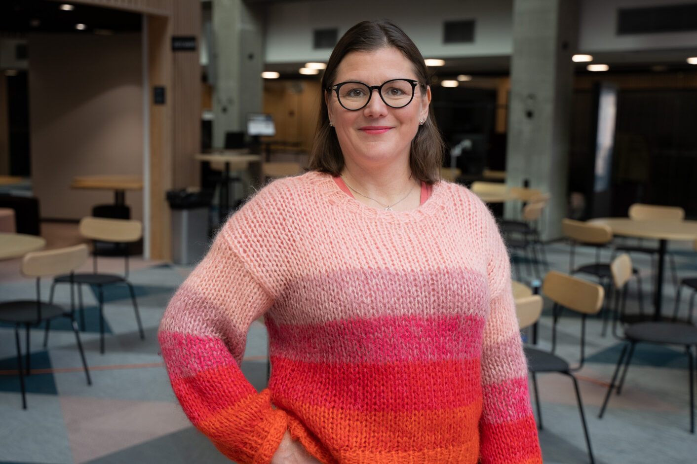 Punaraidalliseen villapaitaan sonustautunut nainen seisoo kameran edessä käsi lantiolla. 