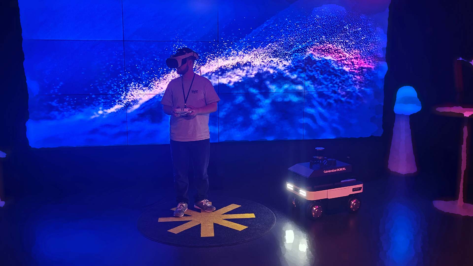 Henkilö virtuaalilasit silmillä, taustalla heijastettuna vesiaihe, henkilön oikealla puolella pieni pyörillä kulkeva robotti