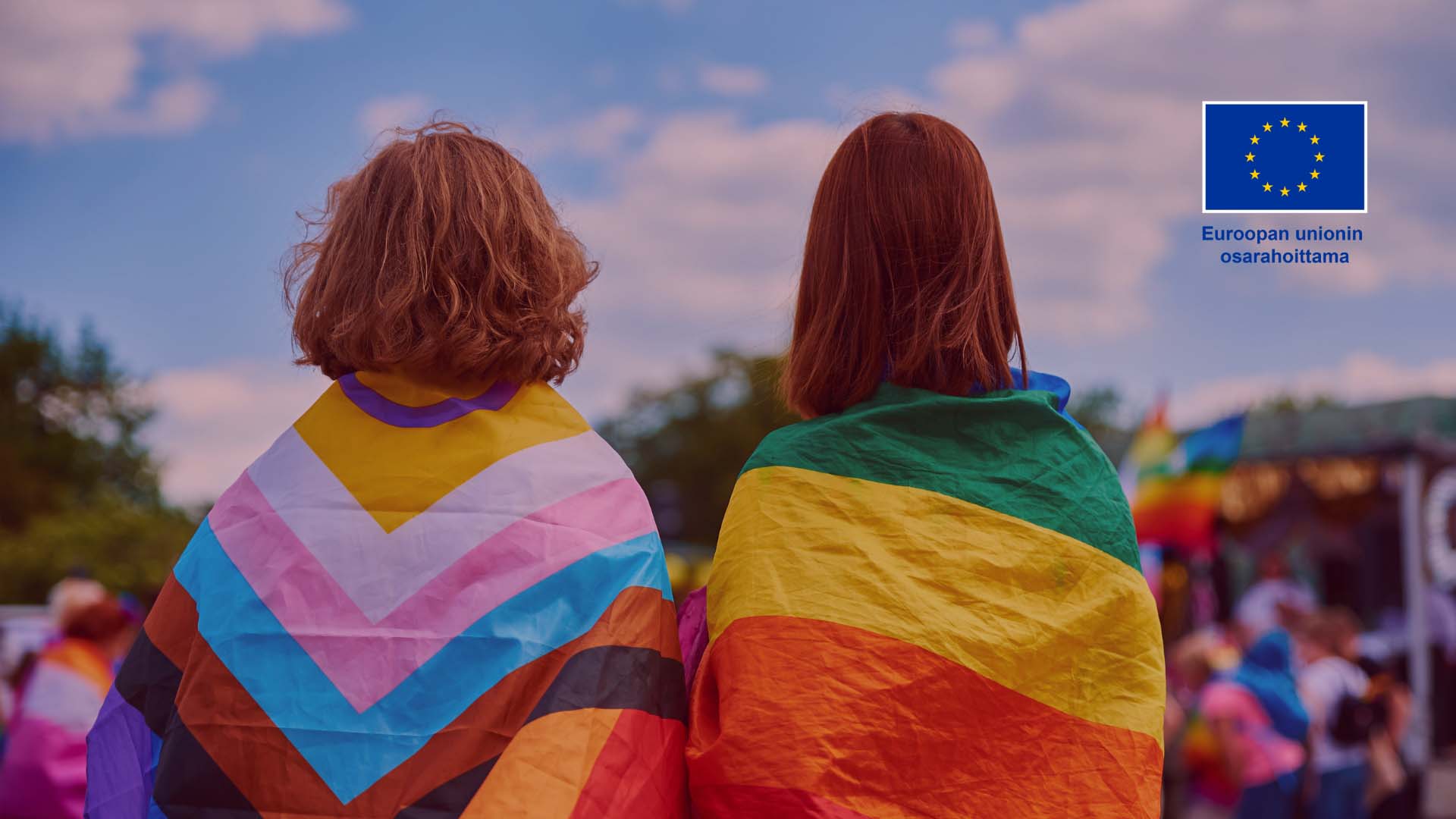 Kaksi henkilöä selin sateenkaariliput harteillaan