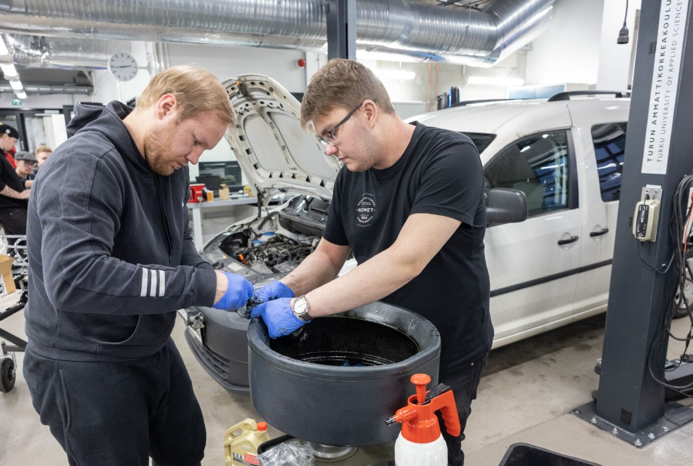 Kaksi henkilö pitelevät mustaa esinettä käsissään, taustalla on auto jonka konepelti on auki.