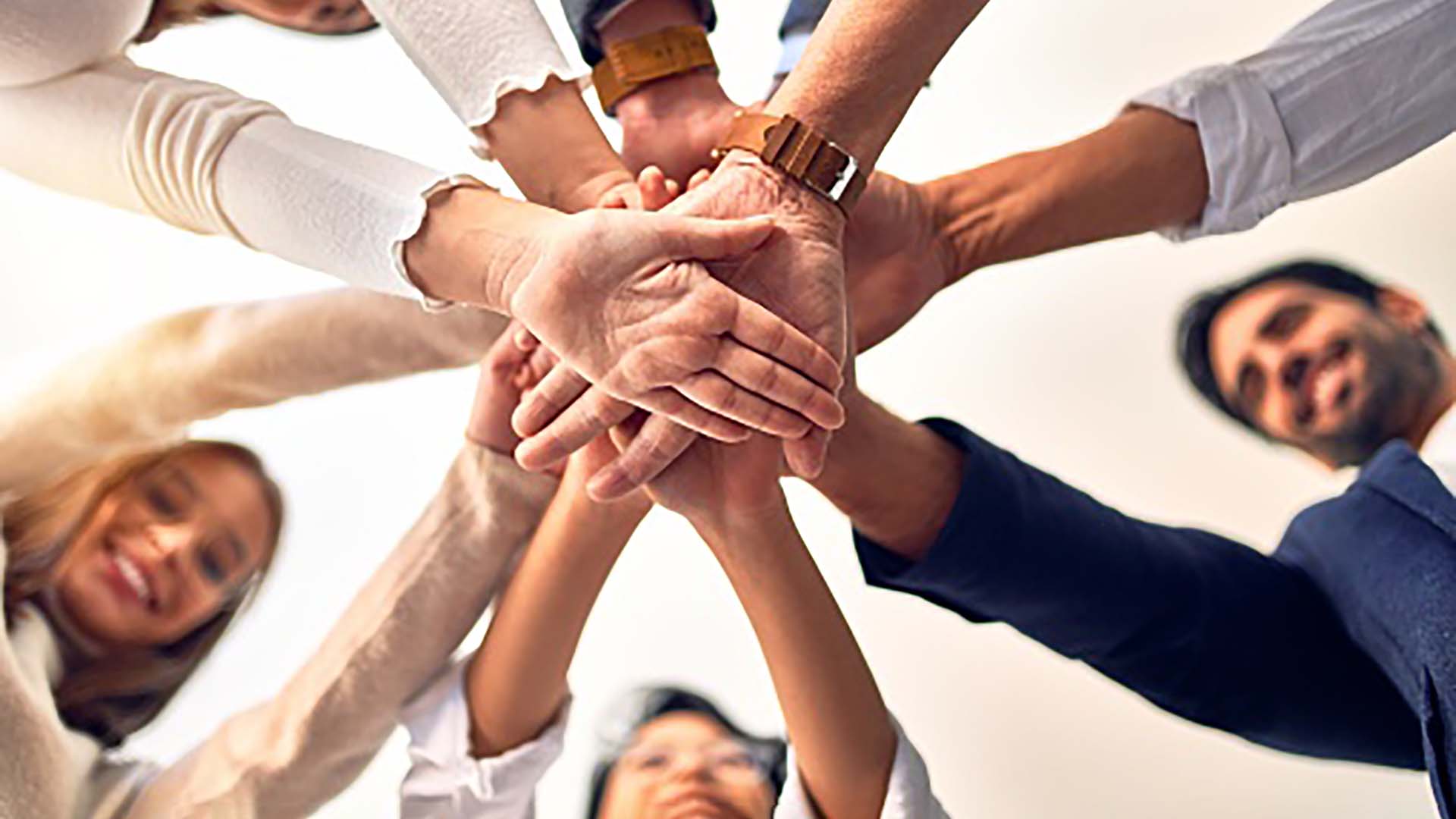 Käsiä päällekkäin pinossa kämmenet alaspäin, myös muutamia kasvoja näkyy taustalla