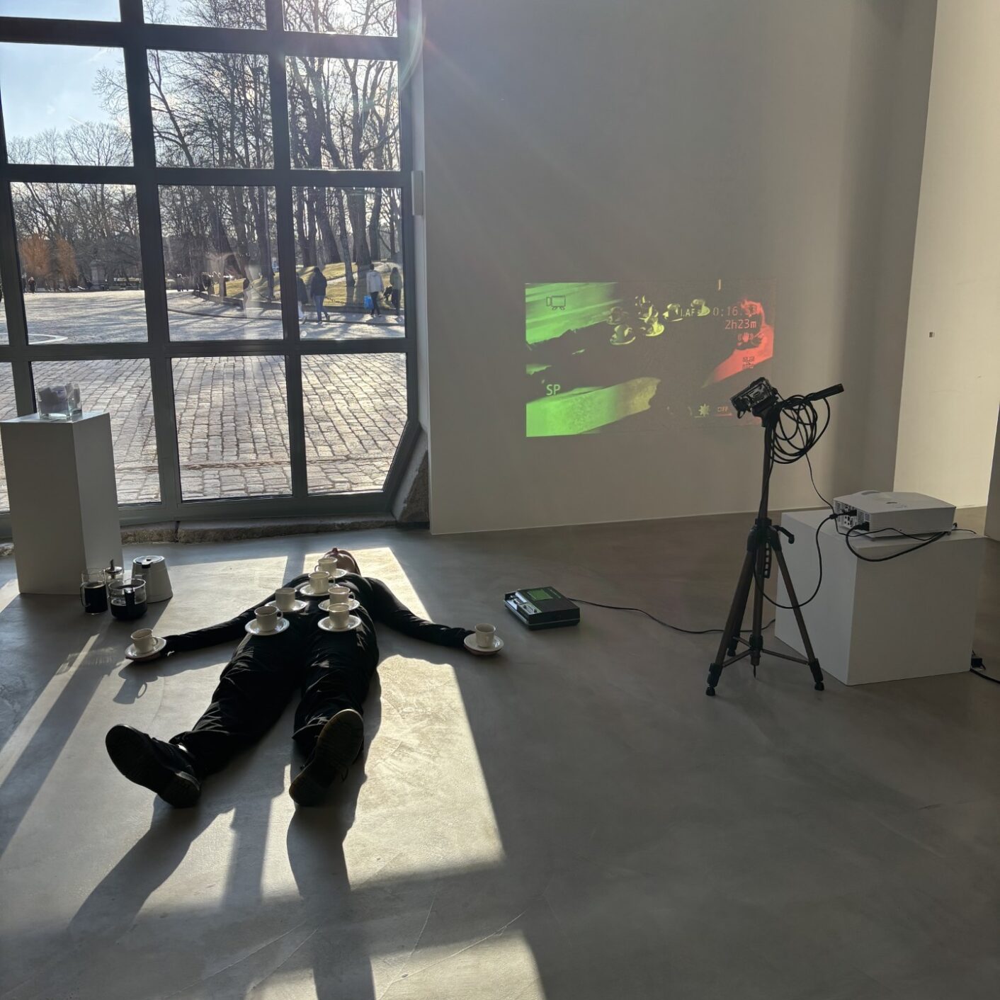 Minea Lehtisen performanssiteoksesta. Kuvassa henkilö lattialla kahvikuppeja vartalonsa päällä, vieressä kasettisoitin sekä projektori, joka heijastaa henkilön kuvan seinälle.