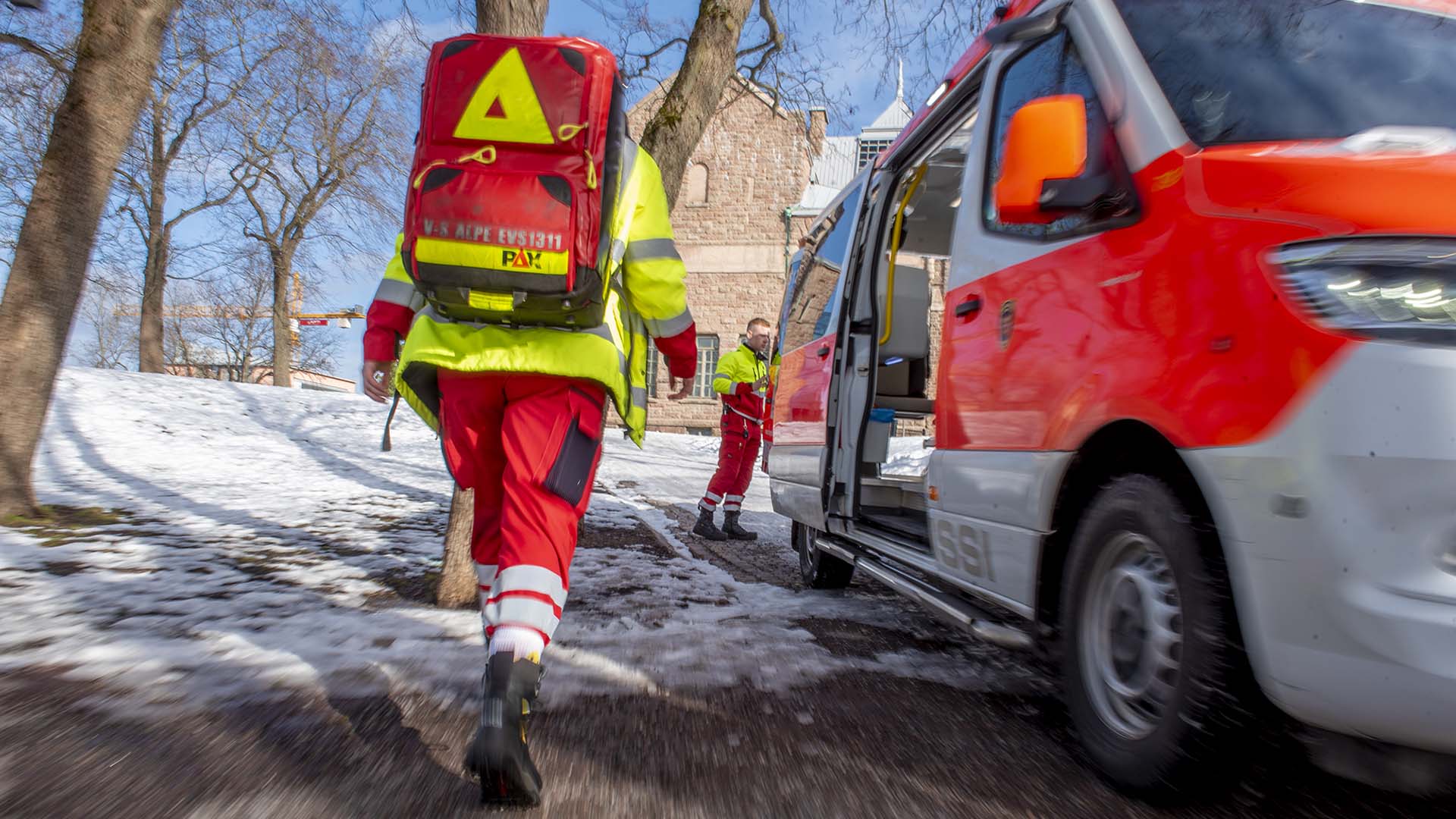 Ensihoitajat menossa työtehtävälle, takana näkyy vanha kivinen rakennus. Etualalla ambulanssi ja ensihoitaja takaa päin
