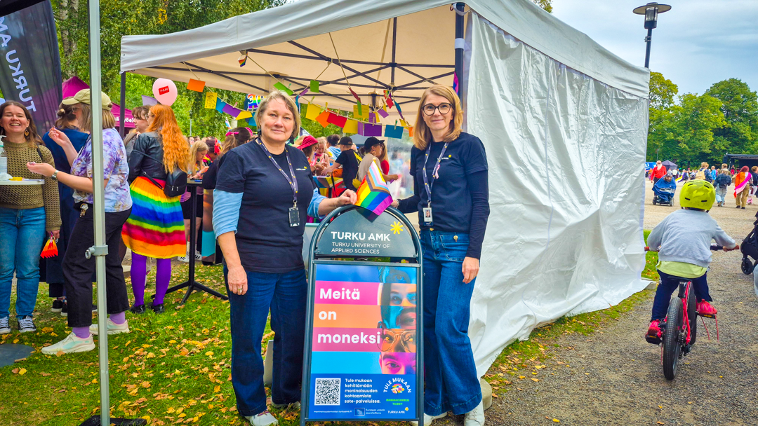 Turku AMK:n Satu Halonen ja Tiina Murto Turku Pride -tapahtumassa, välissä kyltti "Meitä on moneksi".